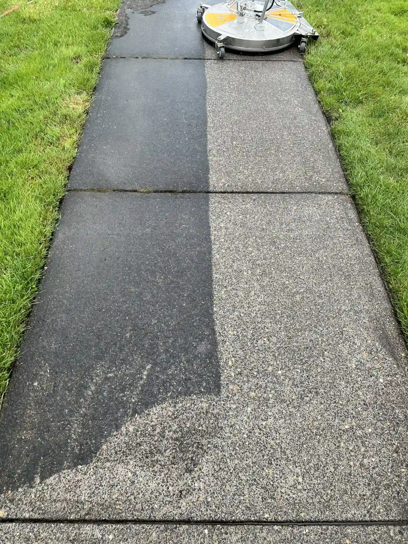 Sidewalk half black with grime, half freshly cleaned, the surface cleaner still in frame